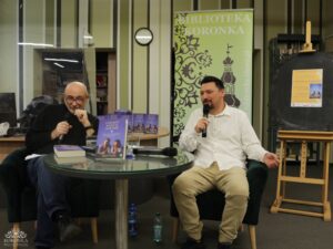 Opis wygenerowany automatycznie: Dwóch mężczyzn siedzi przy okrągłym szklanym stole. Mężczyzna po prawej, ubrany w białą koszulę, mówi do mikrofonu. Mężczyzna po lewej, w okularach i ciemnym swetrze, również trzyma mikrofon. Na stole leżą książki. W tle widoczny jest zielony baner i sztaluga z tablicą.