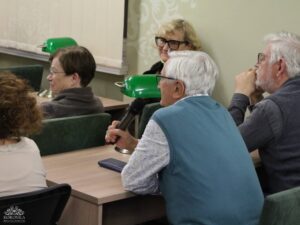 Opis wygenerowany automatycznie: Grupa osób siedzi przy stole w pomieszczeniu. Starszy mężczyzna w niebieskiej kamizelce trzyma mikrofon i wydaje się mówić. Obok niego siedzi inny starszy mężczyzna z brodą, słuchający uważnie. Za mężczyzną z mikrofonem uśmiecha się starsza kobieta w okularach. Na stole widać zieloną lampkę biurkową.