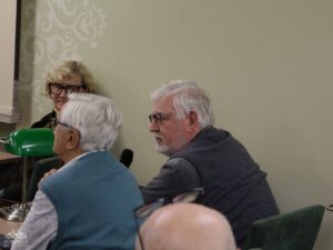 Opis wygenerowany automatycznie: Zbliżenie na trzy osoby siedzące przy stole. Starszy mężczyzna w niebieskiej kamizelce jest widoczny od tyłu, a obok niego siedzi starszy mężczyzna z brodą, patrzący w lewo. Za nimi uśmiecha się starsza kobieta w okularach. W tle jasna ściana z delikatnym wzorem.