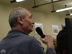 Opis wygenerowany automatycznie: Zbliżenie na starszego mężczyznę z siwymi włosami i wąsami, ubranego w szary sweter, trzymającego czarny mikrofon przy ustach. Ma zamknięte oczy i lekko się uśmiecha, co sugeruje, że śpiewa lub mówi z zaangażowaniem. W tle widać jasną ścianę z obrazami i oświetleniem.