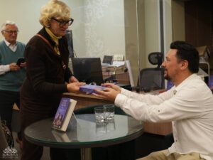 Opis wygenerowany automatycznie: Kobieta w okularach i brązowym swetrze podaje książkę mężczyźnie w białej koszuli, który siedzi przy okrągłym szklanym stole. Na stole leży inna książka. W tle widać starszego mężczyznę.