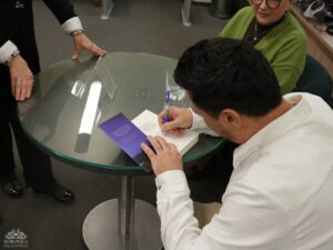 Opis wygenerowany automatycznie: Mężczyzna w białej koszuli siedzi przy okrągłym szklanym stole i podpisuje książkę długopisem. Jego lewa ręka trzyma fioletową zakładkę lub kartę. Obok stołu stoi częściowo widoczna osoba, a za stołem siedzi inna osoba w zielonym swetrze.