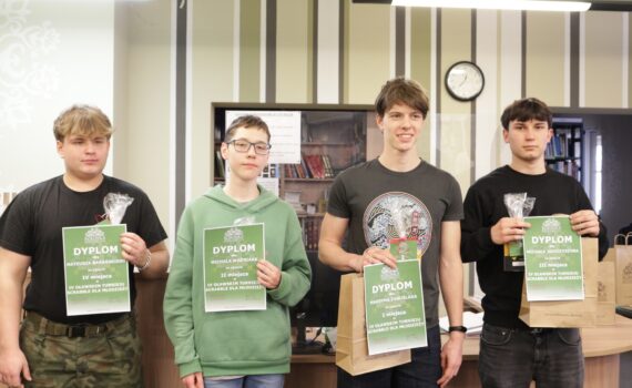Czterech młodych uczestników stoi w bibliotece, trzymając dyplomy i upominki za zajęcie miejsc w turnieju Scrabble dla młodzieży. Wszyscy patrzą w stronę aparatu i uśmiechają się lub stoją poważnie. Za nimi widać półki z książkami oraz zegar na ścianie.