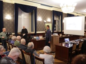 Opis wygenerowany automatycznie: Mężczyzna w ciemnej koszuli przemawia do mikrofonu przy mównicy, obok niego stoi inny mężczyzna. Przed nimi siedzi publiczność, a w tle widoczny jest ekran projektora z wyświetloną grafiką oraz baner z napisem "BIBLIOTEKA KORONKA".