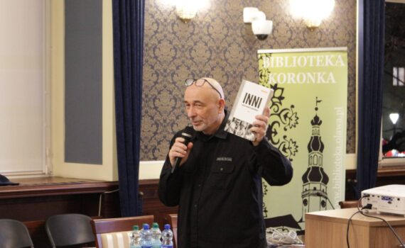 Opis wygenerowany automatycznie: Mężczyzna w okularach i czarnej koszuli trzyma w jednej ręce mikrofon, a w drugiej książkę z napisem "INNI". Stoi przed banerem z logo "BIBLIOTEKA KORONKA" i adresem strony internetowej.