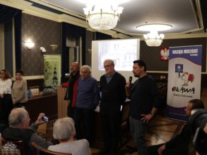 Opis wygenerowany automatycznie: Czterech mężczyzn stoi przed ekranem projektora, na którym wyświetlony jest obraz budynku, oraz przed banerem "URZĄD MIEJSKI W OŁAWIE" z logo koguta. Mężczyzna po prawej stronie, ubrany w ciemną bluzę, trzyma mikrofon i przemawia. Na pierwszym planie widać część publiczności, z której jedna osoba robi zdjęcie telefonem.