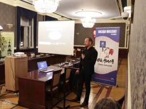 Opis wygenerowany automatycznie: Mężczyzna w czarnym garniturze stoi i przemawia do mikrofonu, prezentując coś na ekranie projektora, na którym widoczna jest prezentacja. Obok niego stoi baner "URZĄD MIEJSKI W OŁAWIE" z logo koguta. Na stole przed ekranem leży otwarty laptop. W tle widoczne są żyrandole, a w prawym dolnym rogu fragment publiczności.