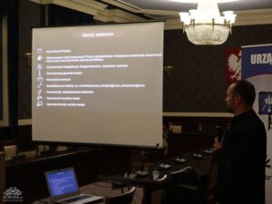 Opis wygenerowany automatycznie: Mężczyzna w ciemnym ubraniu stoi przed ekranem projektora w eleganckiej sali konferencyjnej i prezentuje. Na ekranie widoczny jest slajd z nagłówkiem "Metody badawcze" i listą punktów w języku polskim, m.in. "Kwerenda archiwalna", "Nadzorowanie robót budowlanych", "Dokumentacja fotograficzna", "Konsultacje naukowe" i "Upowszechnienie wyników badań". Na stole przed nim leży otwarty laptop.