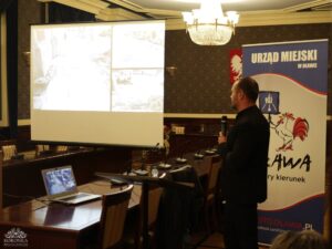 Opis wygenerowany automatycznie: Mężczyzna w ciemnym ubraniu stoi przed ekranem projektora w sali konferencyjnej, trzymając mikrofon. Na ekranie wyświetlone są cztery jasne obrazy, prawdopodobnie przedstawiające architekturę lub krajobraz miejski. Po prawej stronie widoczny jest pionowy baner z napisem "URZĄD MIEJSKI W OŁAWIE" i herbem miasta.