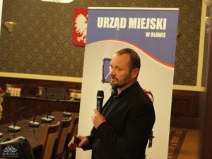 Opis wygenerowany automatycznie: Mężczyzna z brodą, ubrany w ciemną marynarkę, stoi i mówi do mikrofonu. Za nim znajduje się baner z napisem "URZĄD MIEJSKI W OŁAWIE" i godło Polski. W tle widać stoły i krzesła.