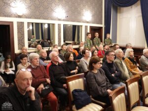 Opis wygenerowany automatycznie: Grupa ludzi siedzi w rzędach, tworząc publiczność na wydarzeniu. W tle widać ozdobne tapety i lustra. W prawym tylnym rogu stoją trzej młodzi mężczyźni w mundurach harcerskich.