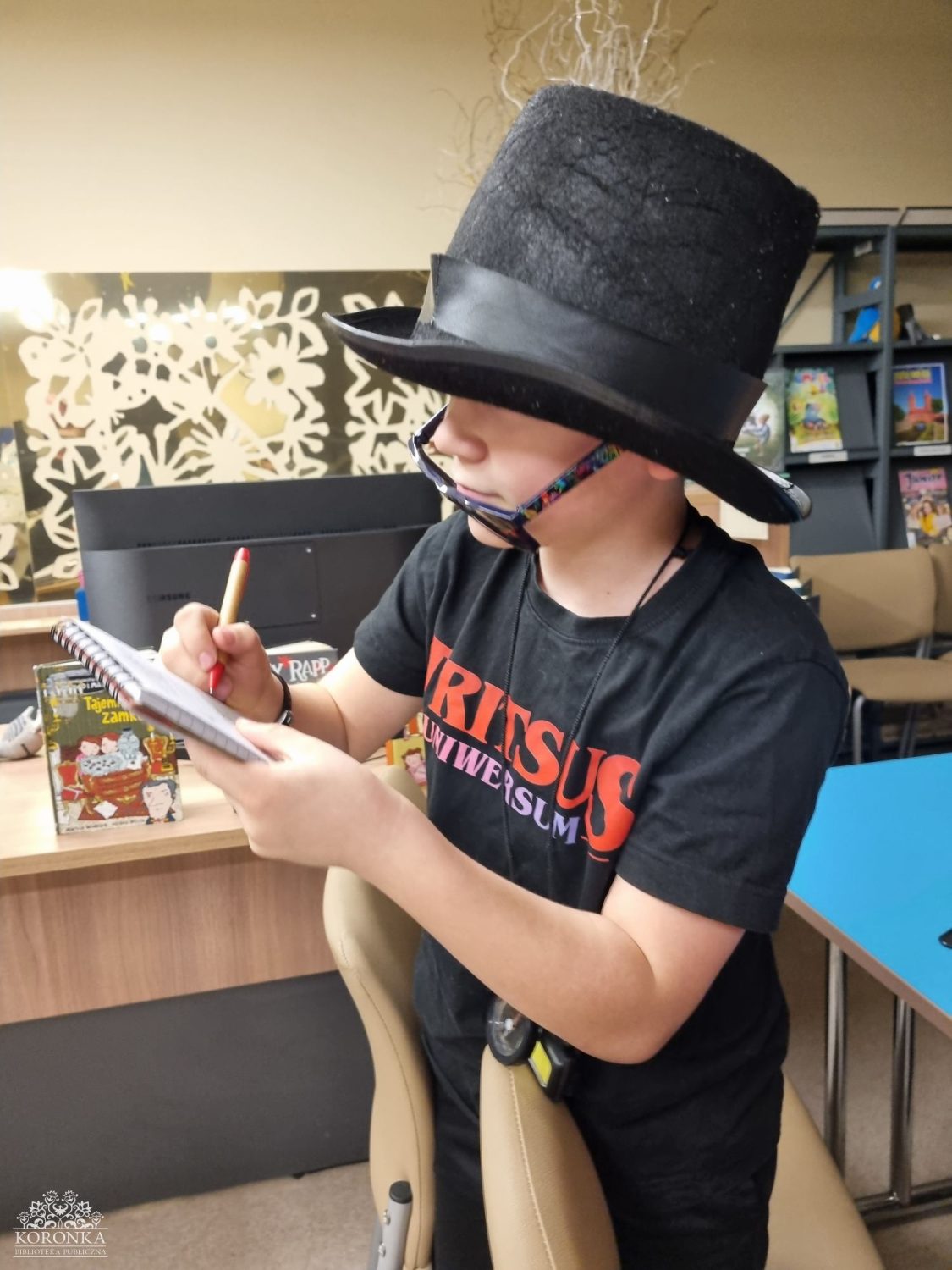 Opis wygenerowany automatycznie: Dziecko w dużym czarnym cylindrze i zsuniętych okularach pisze coś w notesie, siedząc przy biurku w pomieszczeniu przypominającym bibliotekę lub czytelnię.