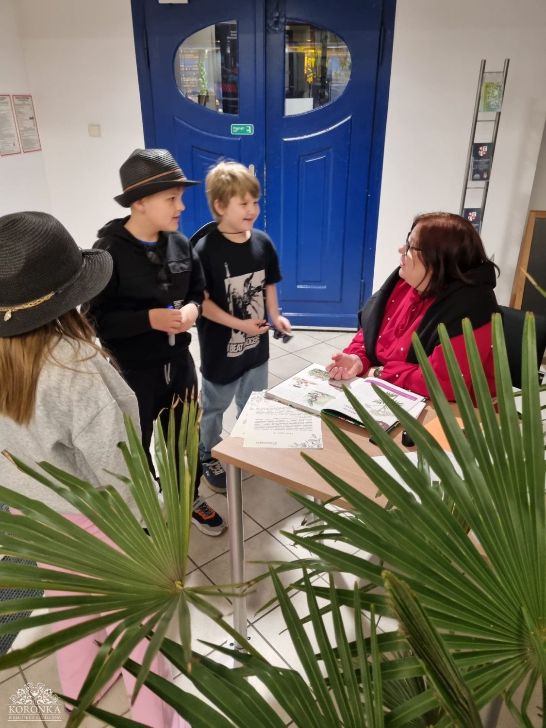 Opis wygenerowany automatycznie: Grupa dzieci rozmawia z kobietą siedzącą przy stole przy wejściu; na stole leżą książki i dokumenty, a na pierwszym planie widoczne są liście rośliny.