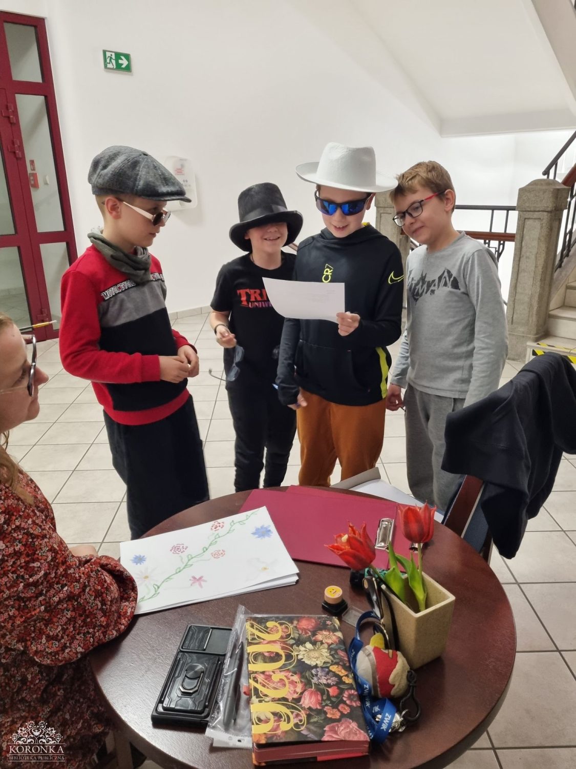 Grupa chłopców przy stoliku Opis wygenerowany automatycznie: Czworo dzieci w wieku szkolnym stoi wokół okrągłego stolika w jasnym holu. Trzech chłopców ma na sobie fantazyjne nakrycia głowy: kaszkiet, czarny cylinder i biały kapelusz typu fedora oraz okulary przeciwsłoneczne. Wspólnie czytają kartkę papieru. Na stole leżą kolorowe rysunki, kwiaty w wazonie i ozdobny kalendarz na rok 2026. Z lewej strony widać fragment siedzącej dorosłej osoby.