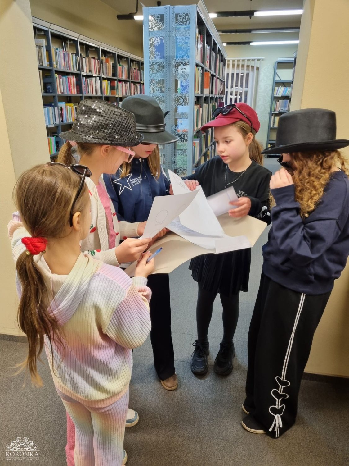 Grupa dziewczynek w bibliotece Opis wygenerowany automatycznie: Pięć dziewczynek stoi w przejściu między regałami bibliotecznymi, tworząc ciasne koło. Większość z nich ma na sobie przebrania: błyszczące kapelusze, czarny cylinder oraz okulary przeciwsłoneczne. Dzieci z dużym zaangażowaniem przeglądają plik dokumentów w białej teczce. W tle widać rzędy książek na półkach.