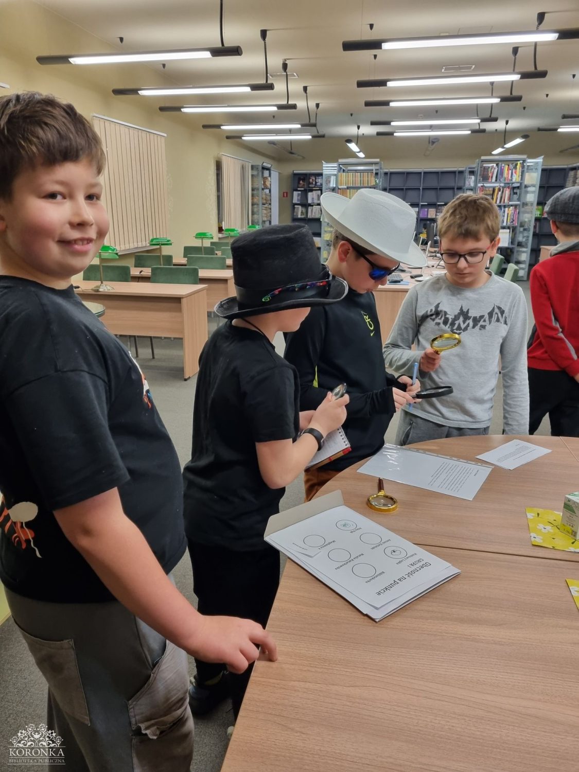 Mali detektywi w czytelni Opis wygenerowany automatycznie: Grupa chłopców w przebraniach (kapelusze, okulary przeciwsłoneczne) bawi się w detektywów w nowoczesnej bibliotece. Dwóch chłopców używa lup do badania dokumentów rozłożonych na drewnianym stole. Na pierwszym planie po lewej stronie uśmiechnięty chłopiec w czarnym t-shircie patrzy w stronę obiektywu. W tle widoczne są biurka do pracy i regały z książkami.