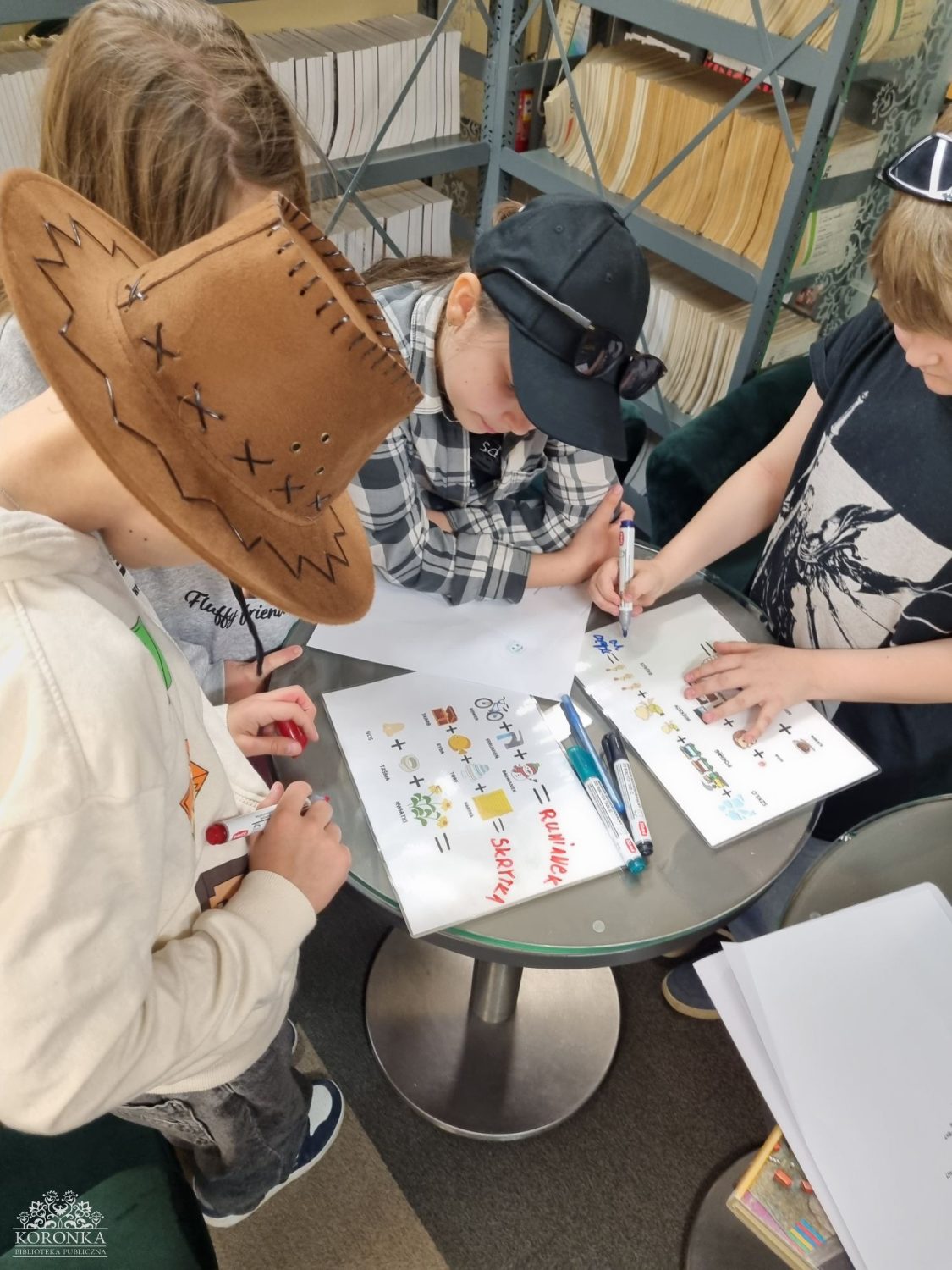 Praca zespołowa przy zagadce Opis wygenerowany automatycznie: Widok z góry na grupę trojga dzieci zgromadzonych wokół małego, szklanego stolika. Chłopiec po lewej nosi brązowy kapelusz kowbojski. Pośrodku dziewczynka w bejsbolówce i koszuli w kratę pisze coś flamastrem. Po prawej inny chłopiec wskazuje na kartkę. Na stole leżą dwa arkusze papieru z rebusem obrazkowym i hasłami napisanymi flamastrem: „RUMIANEK” i „SKRYTKA”. W tle widać biblioteczne regały.