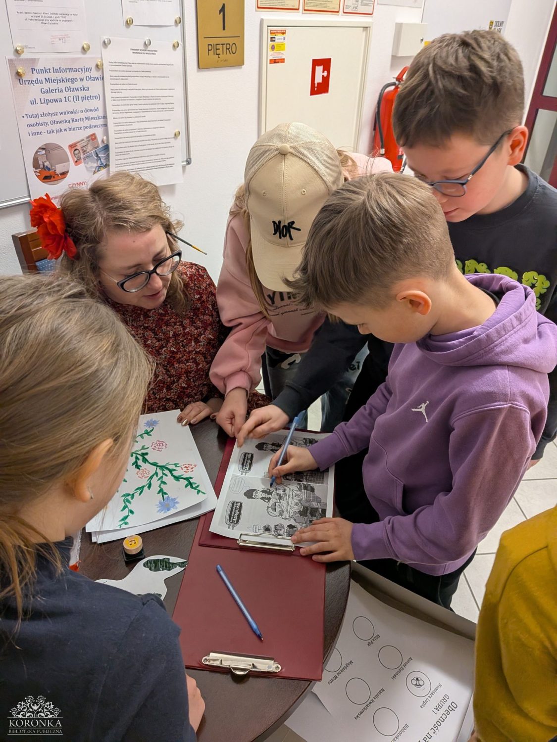 Warsztaty przy stoliku Opis wygenerowany automatycznie: Widok z góry na czworo dzieci i bibliotekarkę w okularach z czerwoną ozdobą we włosach, skupionych wokół biurka. Dwoje dzieci po prawej stronie wypełnia arkusze z zadaniami umieszczone na podkładkach z klipsem. Jedno dziecko nosi czapkę z daszkiem. Przed bibliotekarką leży kartka z kolorowym rysunkiem roślin. W tle widać białą ścianę z tablicą informacyjną, gaśnicą i tabliczką „1 PIĘTRO”.