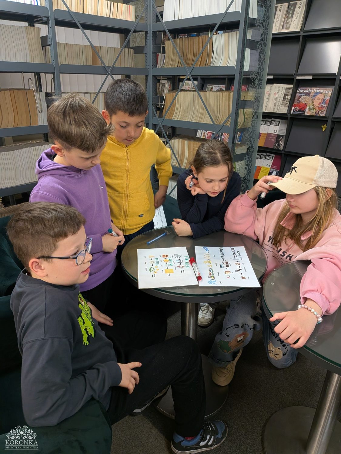 Rozwiązywanie rebusów w bibliotece Opis wygenerowany automatycznie: Pięcioro dzieci stoi lub siedzi wokół okrągłego stolika w bibliotece. Uważnie przyglądają się dwóm kartkom z rebusami obrazkowymi. Chłopiec w okularach i ciemnej bluzie siedzi na pierwszym planie, podczas gdy pozostałe dzieci (m.in. w żółtej i fioletowej bluzie z kapturem oraz różowej bluzie i beżowej czapce z daszkiem) pochylają się nad zadaniem. W tle widać regały z czasopismami i książkami.