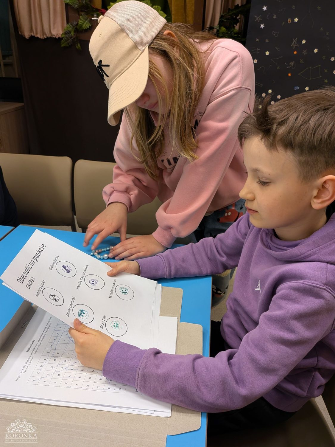 Sprawdzanie listy obecności Opis wygenerowany automatycznie: Zbliżenie na dwoje dzieci przy niebieskim stoliku. Chłopiec w fioletowej bluzie trzyma kartkę z napisem „Obecność na punkcie GRUPA 1”, na której widnieją nazwiska i przypisane do nich puste kółka z małymi grafikami. Obok niego stoi dziewczynka w różowej bluzie i beżowej czapce z daszkiem, która kładzie na stole ozdobną bransoletkę z koralików. Pod spodem widoczny jest arkusz z literową wykreślanką.