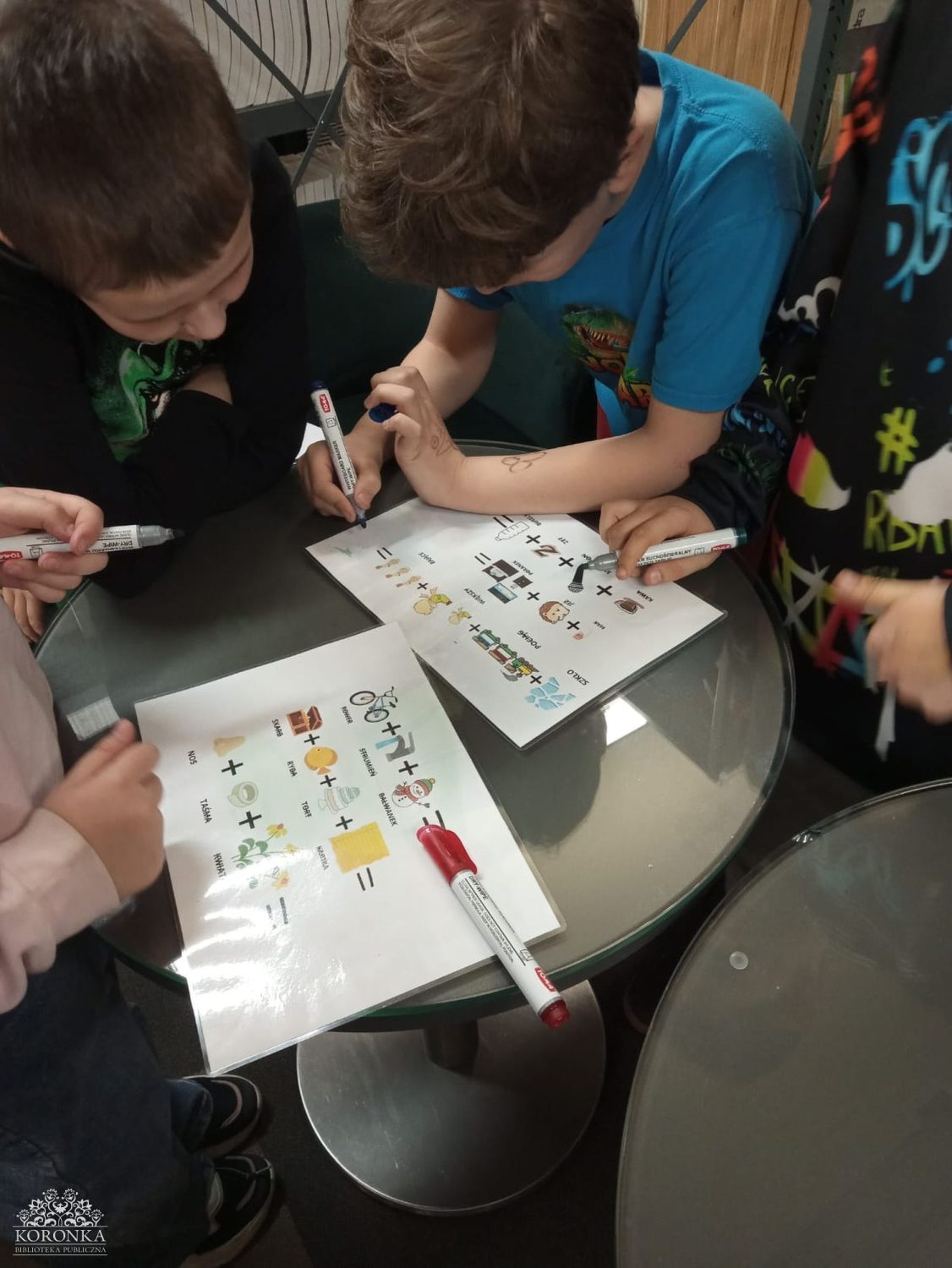 Rozwiązywanie detektywistycznych rebusów Opis wygenerowany automatycznie: Zbliżenie na dłonie trojga dzieci piszących mazakami po zalaminowanych arkuszach z rebusami. Na kartkach widoczne są kolorowe obrazki (rower, bałwanek, ryba) połączone znakami plus, tworzące zagadki słowne. Dzieci pracują wspólnie przy okrągłym, szklanym stoliku w bibliotece.