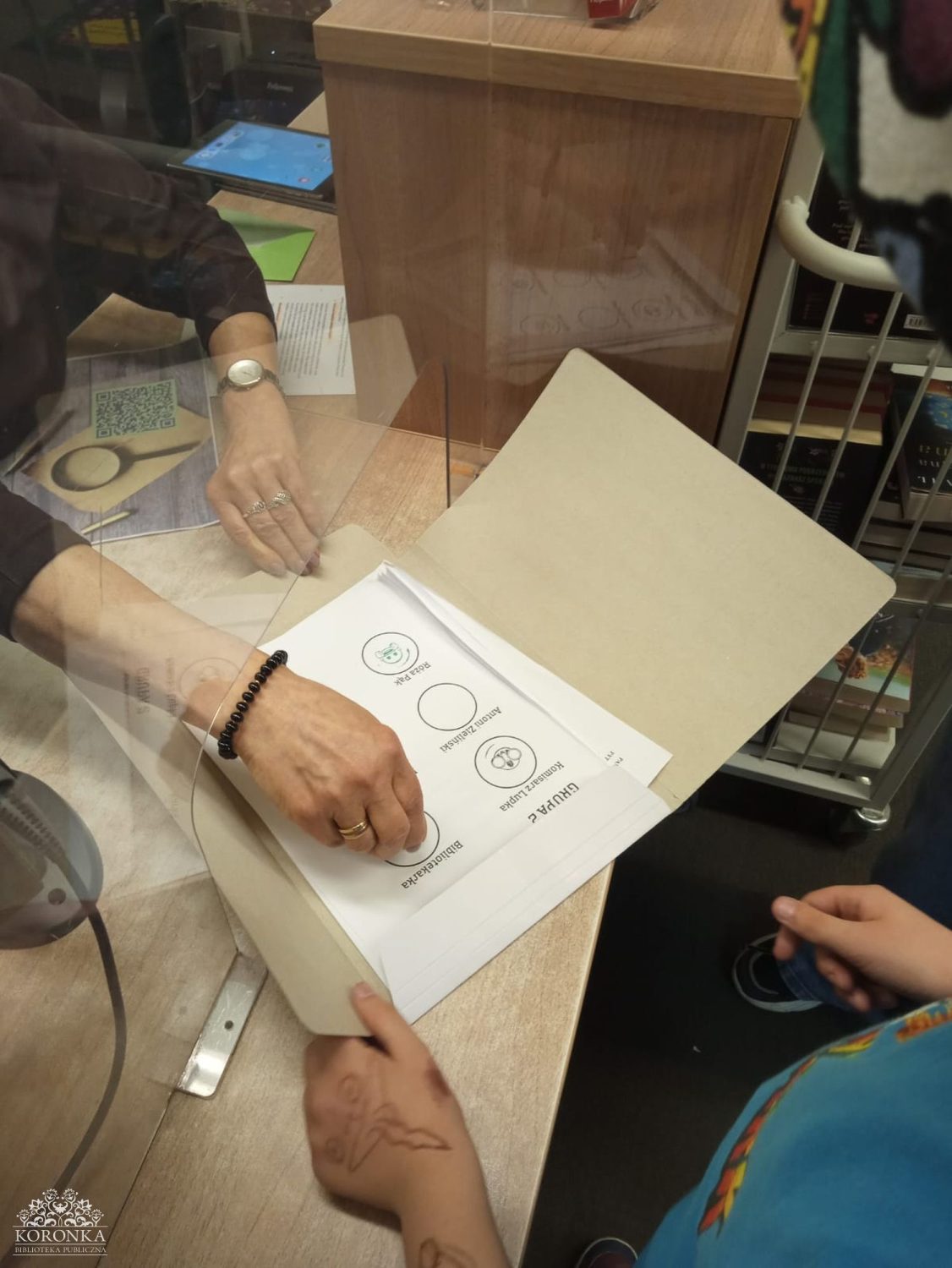 Potwierdzanie zadań w teczce Opis wygenerowany automatycznie: Widok z góry na biurko oddzielone szybą ochronną. Dorosła osoba stawia pieczątkę na liście obecności wewnątrz teczki z napisem „GRUPA 2”. Na liście widnieją nazwiska i rysunkowe ikony. Z dołu widać dłonie dziecka trzymającego teczkę, na których narysowany jest tatuaż z henny lub długopisu przedstawiający motyla.