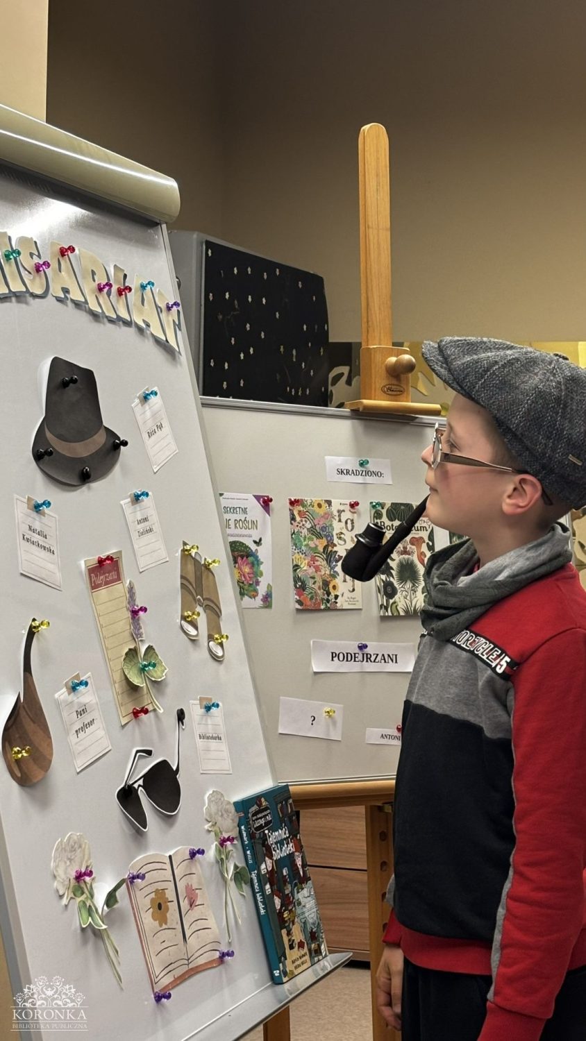 Opis wygenerowany automatycznie: Chłopiec w szarym kaszkiecie, czerwono-szarej bluzie i okularach stoi obok tablicy z napisem "KOMISARIAT". Patrzy w lewo na przypięte do niej elementy, w tym czarny kapelusz, lupy i kartki z napisami. Na tablicy obok widoczne są okładki książek i napisy "SKRADZIONO:" oraz "PODEJRZANI".