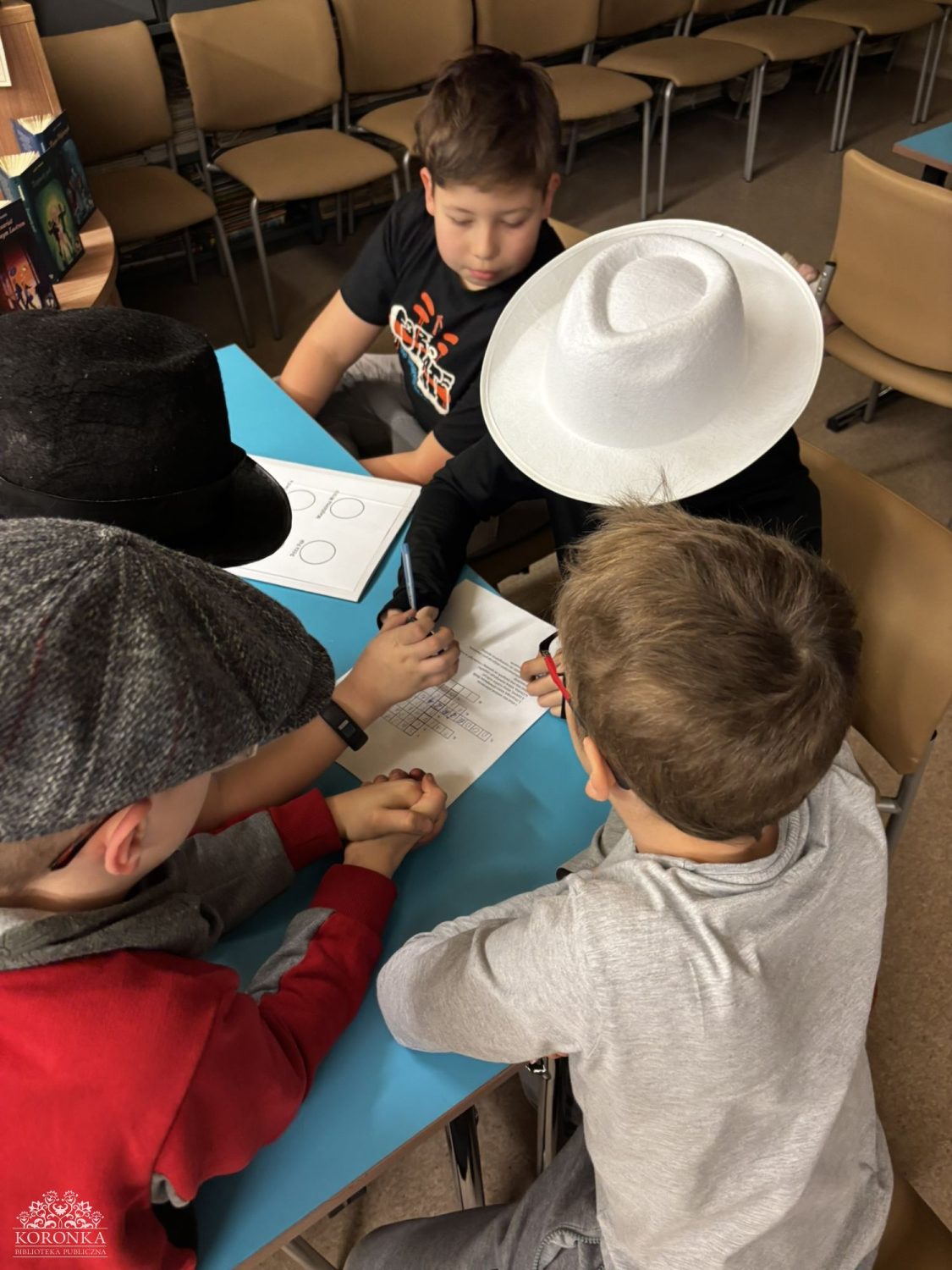 Opis wygenerowany automatycznie: Czterech chłopców siedzi przy niebieskim stole, pochylając się nad kartką papieru. Widać, że jeden z nich pisze długopisem po arkuszu z kratkami. Dwaj chłopcy mają na głowach kapelusze – czarny cylinder i biały kapelusz typu fedora, a trzeci nosi szary kaszkiet. W tle widać rzędy krzeseł.