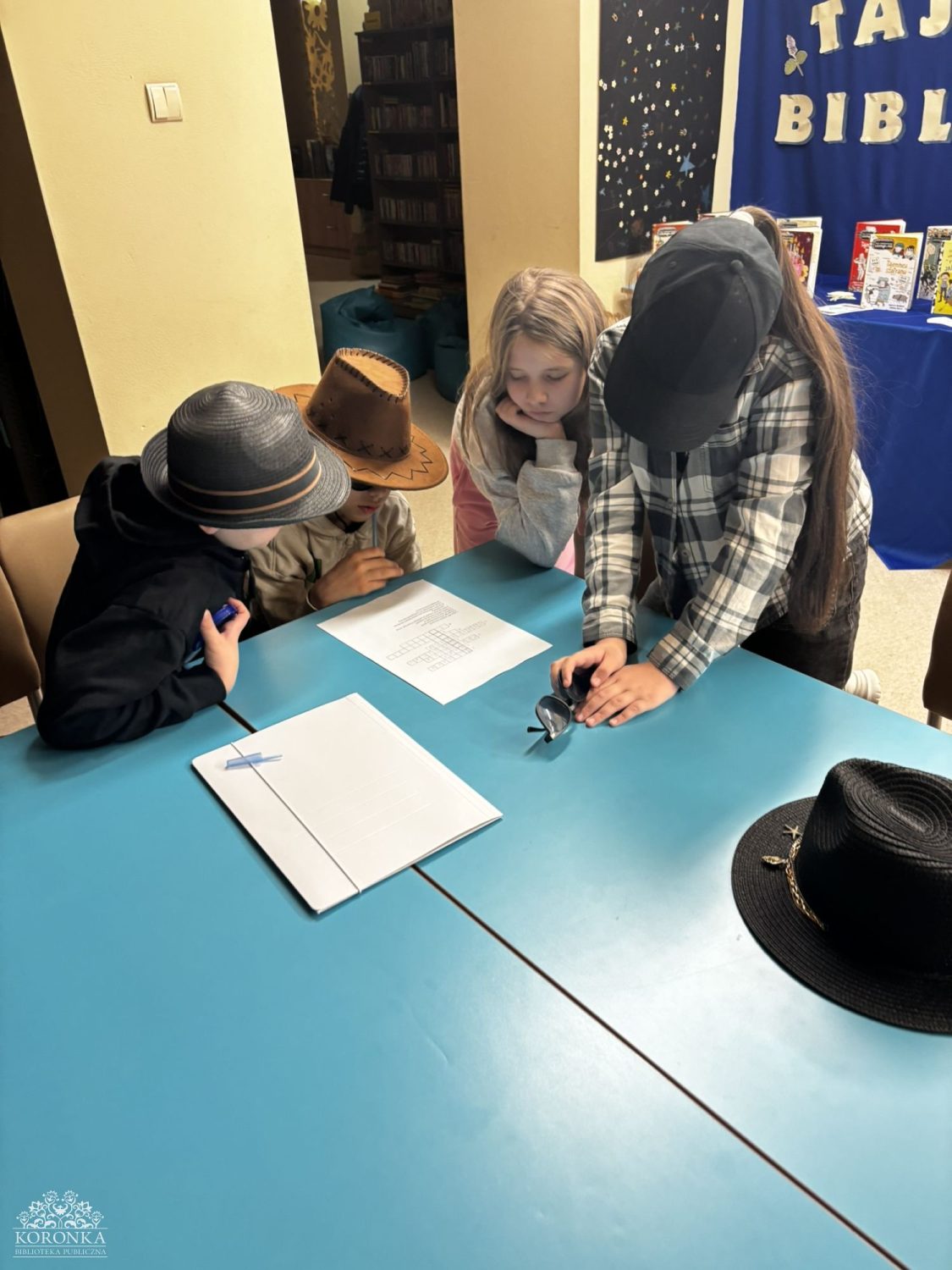 Opis wygenerowany automatycznie: Czworo dzieci stoi wokół niebieskiego stołu w bibliotece. Uważnie przyglądają się kartce z krzyżówką lub innym zadaniem. Jedna z dziewczynek w czarnej bejsbolówce i koszuli w kratę trzyma parę okularów przeciwsłonecznych, a chłopiec w kapeluszu kowbojskim pochyla się nad stołem. Na krawędzi blatu leży czarny kapelusz z rondem. W tle widać regał z książkami i niebieską dekorację.