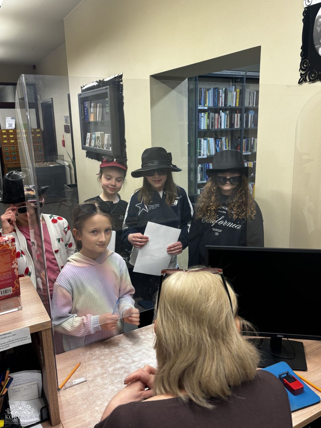 Opis wygenerowany automatycznie: Grupa pięciu dziewczynek w różnorodnych kapeluszach i okularach przeciwsłonecznych stoi przed ladą biblioteczną, oddzielona przezroczystą pleksiglasową osłoną od siedzącej za nią bibliotekarki. Dwie dziewczynki noszą czarne kapelusze i ciemne okulary. Jedna z nich trzyma kartkę papieru. Bibliotekarka siedzi tyłem do aparatu, widoczny jest monitor i przybory biurowe na ladzie. W tle widać regały z książkami.
