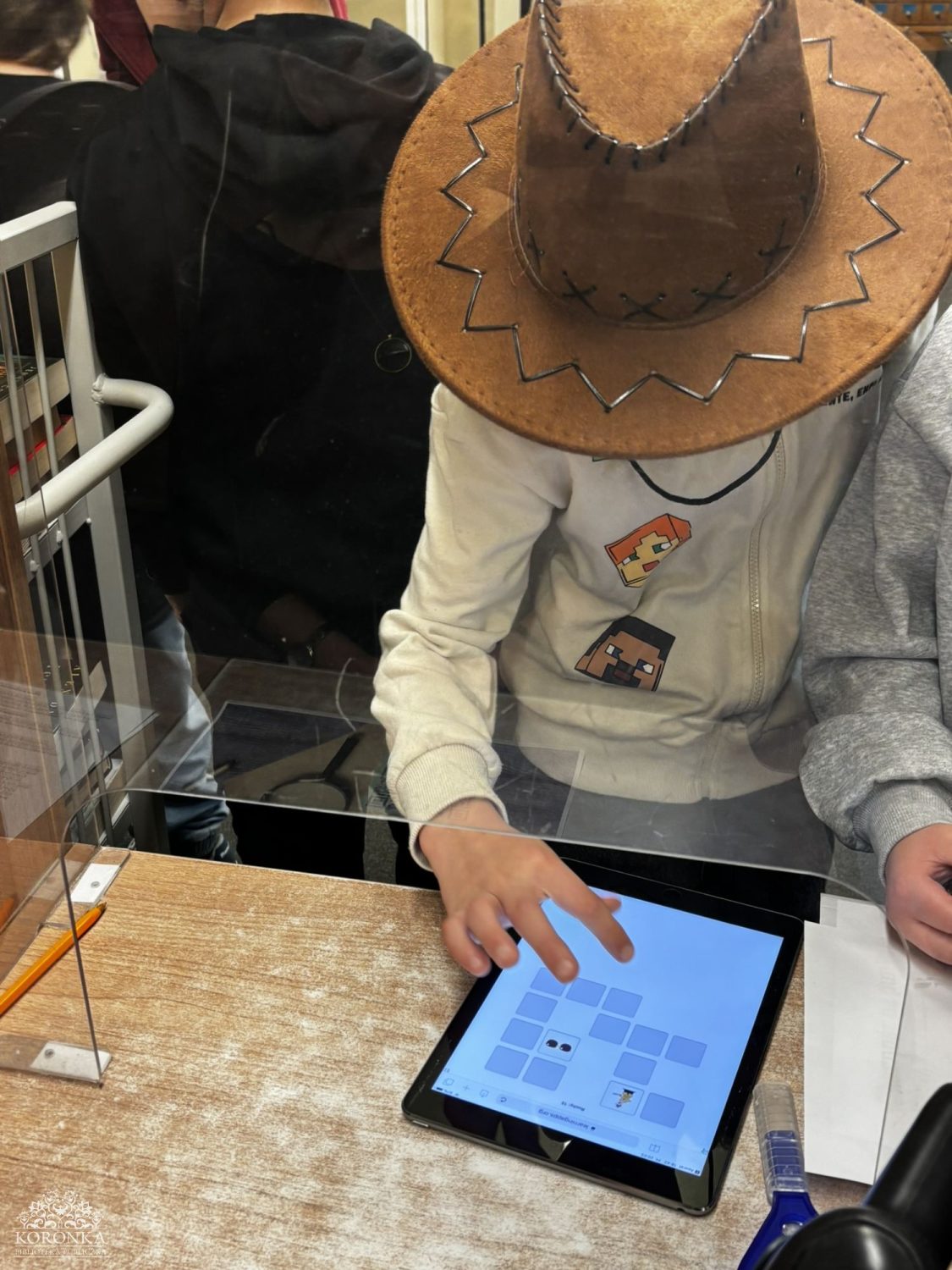 Opis wygenerowany automatycznie: Dziecko w kowbojskim kapeluszu i jasnej bluzie stoi tyłem, patrząc w stronę bibliotekarki i grupy rówieśników siedzących na krzesłach w głębi sali. Spotkanie odbywa się w bibliotece.