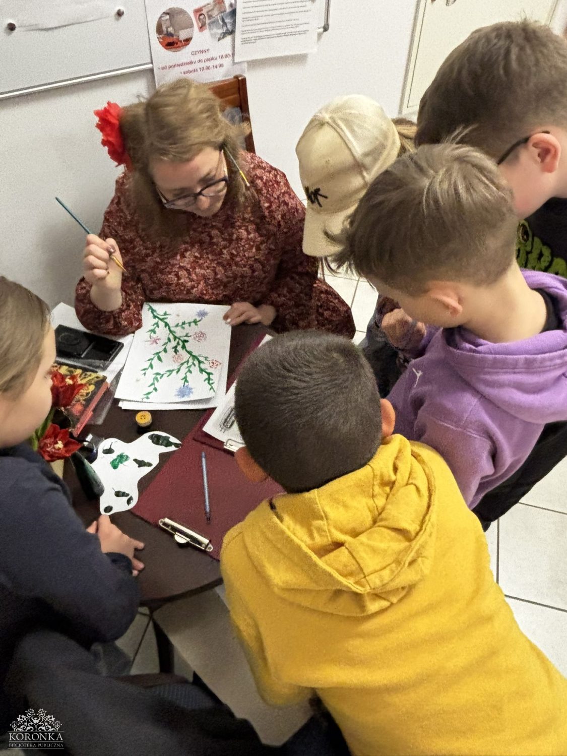 Rozwiązywanie rebusów (zbliżenie) Opis wygenerowany automatycznie: Zbliżenie na dłonie dzieci i bibliotekarki pracujących przy małym stoliku. Wspólnie analizują arkusze z kolorowymi rebusami i obrazkami. Na stole leżą kolorowe pisaki i pieczątka.