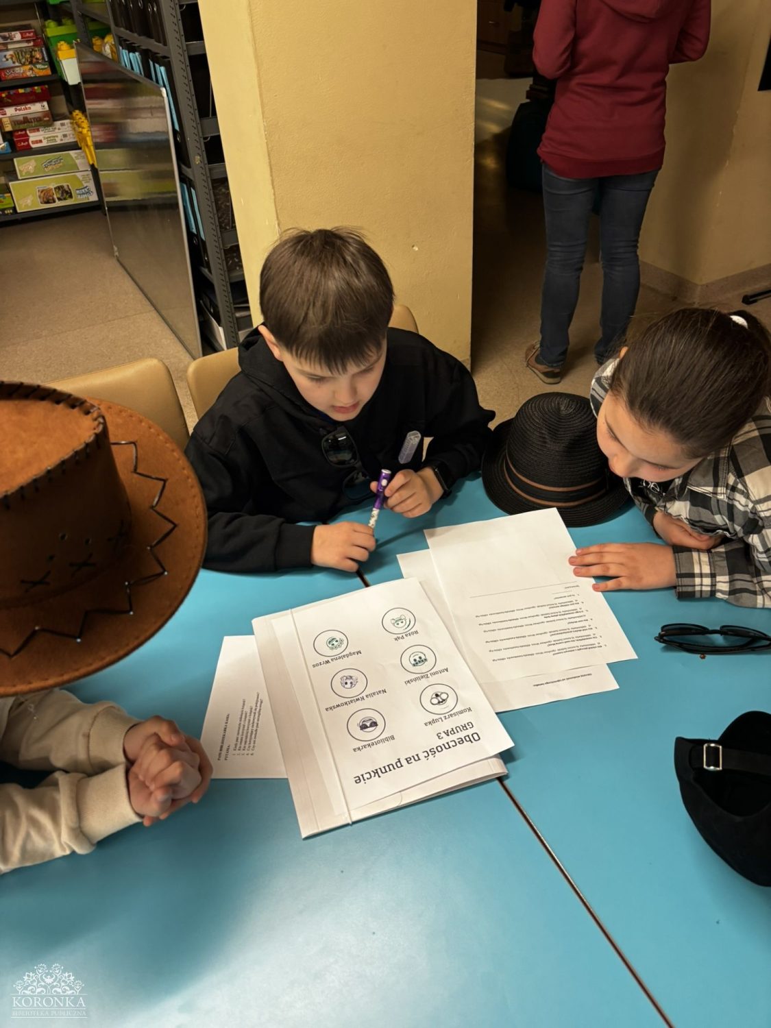 Praca nad krzyżówką Opis wygenerowany automatycznie: Widok z góry na dzieci skupione przy niebieskim stoliku w bibliotece. Wspólnie rozwiązują zadanie W kadrze widoczne są różne nakrycia głowy: kapelusz kowbojski, czapka z daszkiem oraz cylindry.