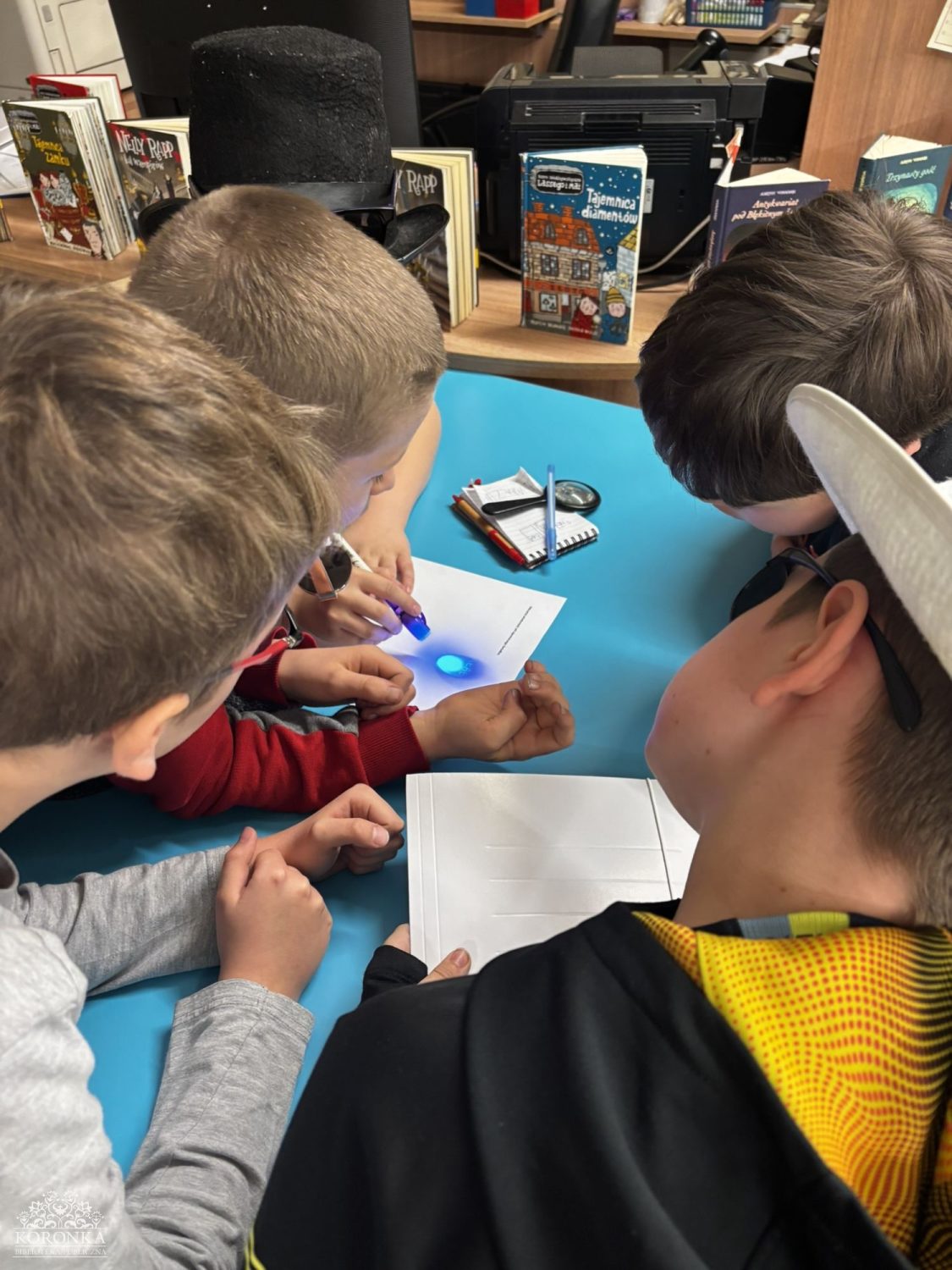 Praca nad krzyżówką Opis wygenerowany automatycznie: Widok z góry na dzieci skupione przy niebieskim stoliku w bibliotece. Wspólnie rozwiązują zadanie