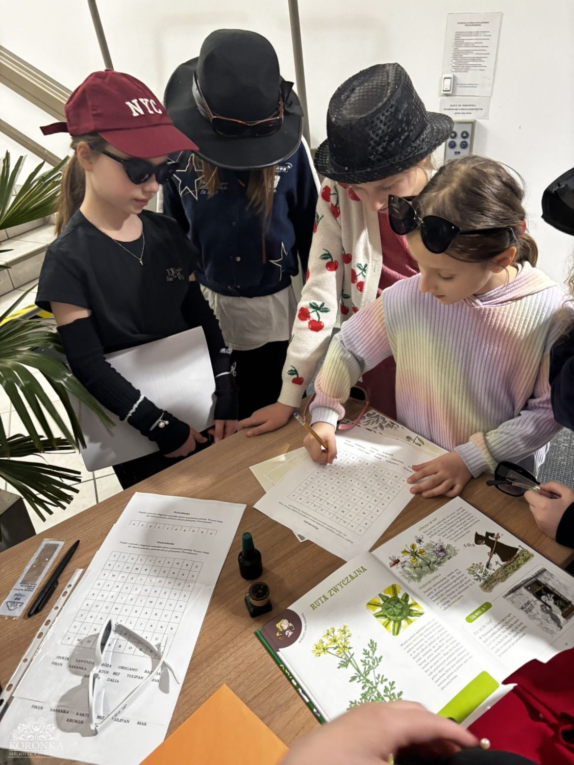 Praca nad krzyżówką Opis wygenerowany automatycznie: Widok z góry na czworo dzieci skupionych przy stoliku w bibliotece. Wspólnie rozwiązują dużą wykreślankę literową wydrukowaną na arkuszu papieru. W kadrze widoczne są różne nakrycia głowy: kapelusz kowbojski, czapka z daszkiem oraz cylindry.