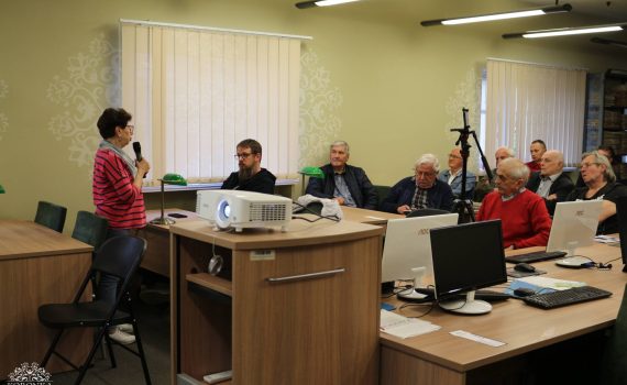 Opis wygenerowany automatycznie: Kobieta w różowym swetrze w paski stoi po lewej stronie sali i mówi do mikrofonu, zwracając się do zgromadzonej publiczności. Przy stołach komputerowych siedzi grupa głównie starszych osób, w tym mężczyzna w czerwonym swetrze, słuchających wypowiedzi. Na środku sali, na podwyższeniu, stoi włączony projektor multimedialny.