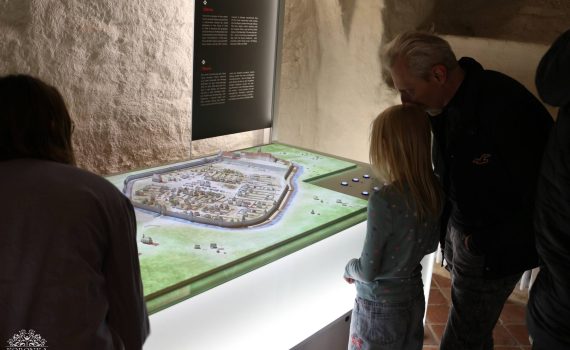 Opis wygenerowany automatycznie: Mężczyzna i dziewczynka z bliska przyglądają się dużej makiety przedstawiającej historyczną zabudowę miasta Oława z murami obronnymi. Makieta jest umieszczona na podświetlanym od dołu stole. Na ścianie obok wisi czarna tablica informacyjna z opisami w języku polskim i niemieckim pod tytułem „Oława”. Po lewej stronie widoczny jest tył głowy innej osoby obserwującej wystawę.