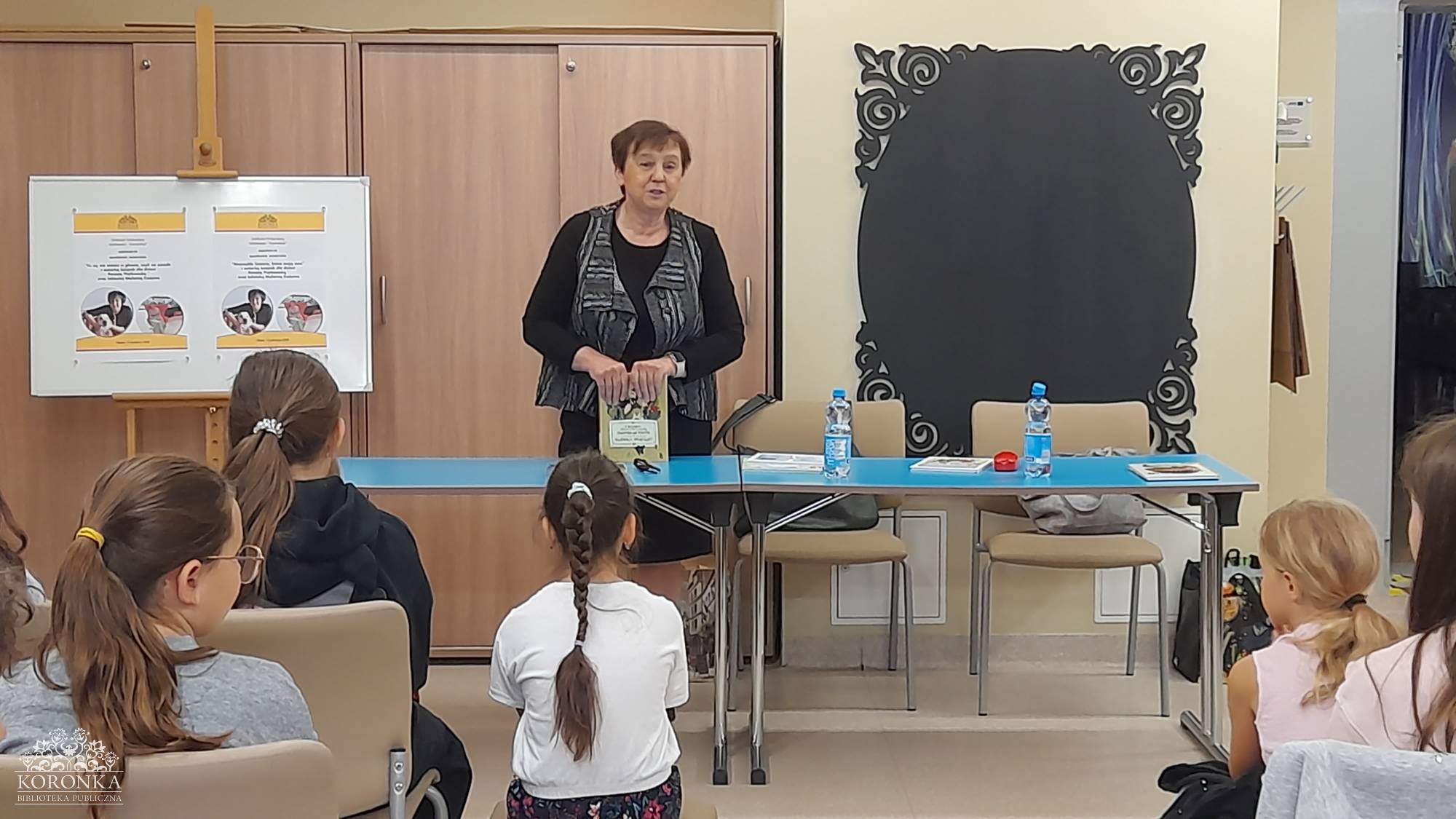 Opis wygenerowany automatycznie: Kobieta w okularach, czarnej bluzce i wzorzystej kamizelce stoi za niebieskim stołem w bibliotece publicznej, trzymając w rękach książkę dla dzieci. Przed nią, tyłem do obiektywu, siedzi grupa dzieci słuchających prezentacji. Po lewej stronie widoczny jest stojak z plakatami informacyjnymi, a w lewym dolnym rogu logo Biblioteki Publicznej „Koronka”.