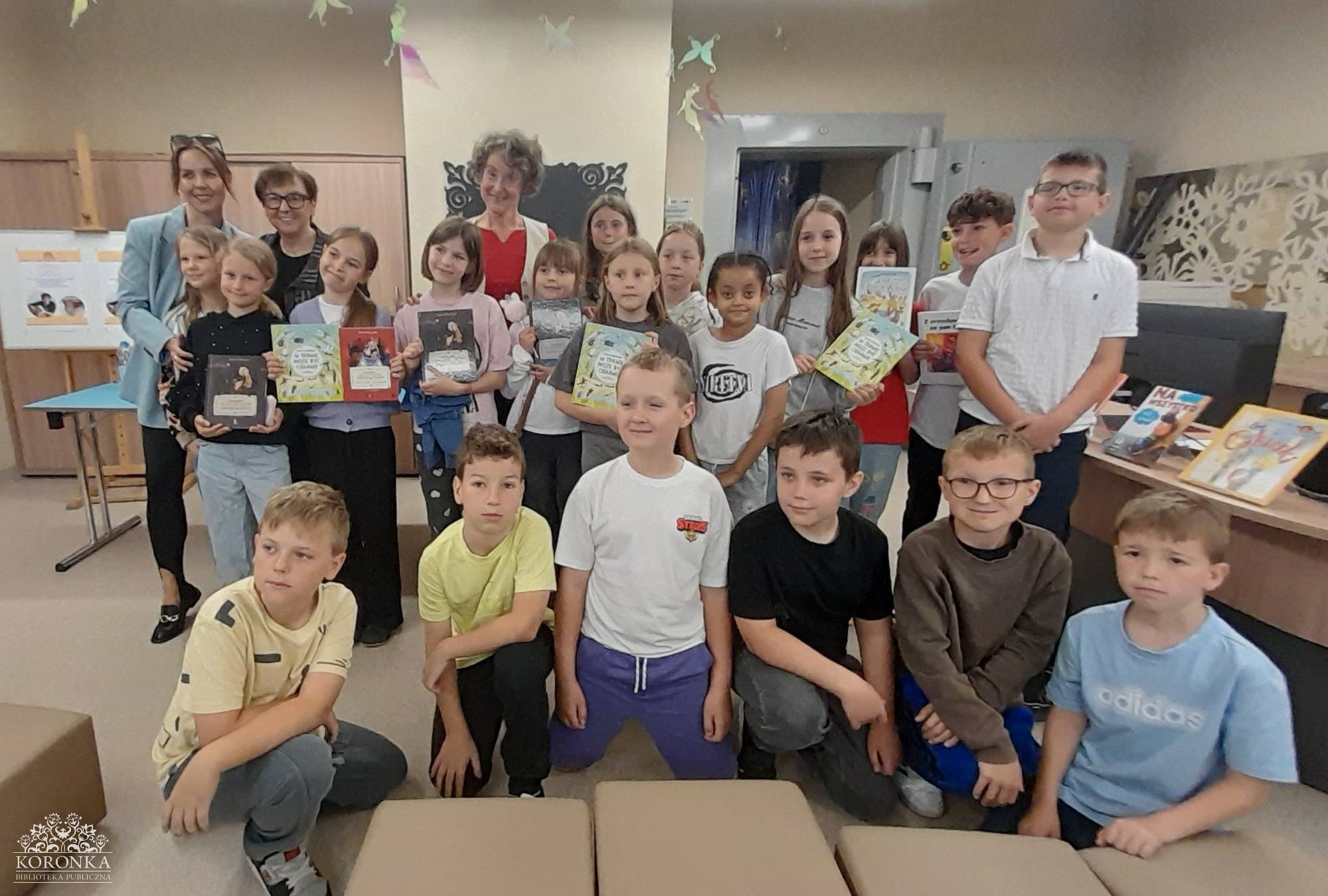 Opis wygenerowany automatycznie: Pozowane zdjęcie grupowe uczestników spotkania w bibliotece. Duża grupa dzieci oraz trzy dorosłe kobiety stoją i klęczą w kilku rzędach, uśmiechając się do aparatu. Wiele dzieci dumnie trzyma w dłoniach egzemplarze nowo zakupionych lub podpisanych książek.