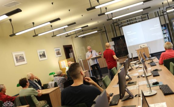 Opis wygenerowany automatycznie: Widok z ukosu na salę wykładową lub komputerową w bibliotece. Wzdłuż długiego stołu z monitorami i laptopami siedzą uczestnicy spotkania. W głębi sali, przy ekranie projekcyjnym, stoi starszy mężczyzna w jasnej koszuli mówiący do mikrofonu. Przed nim tyłem stoi mężczyzna w czerwonej koszulce. Na ekranie wyświetlany jest slajd z napisem „Oława nieoczywista”. W lewym dolnym rogu znajduje się logo „Koronka Biblioteka Publiczna”.